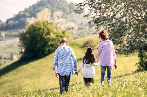 A grandfather, mother, and daughter walk hand in hand across a grassy field towards a beautiful setting sun.