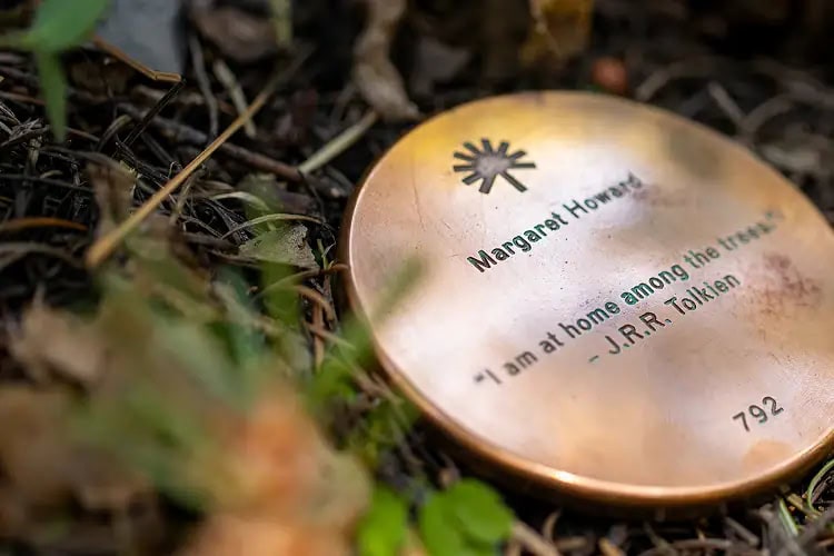 Close-up of a bronze memorial marker at the base of a tree reading ‘Margaret Howard – I am home among the trees,’ symbolizing a lasting tribute in nature.