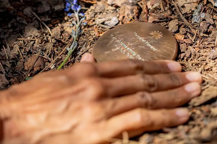 Close-up of a hand reaching toward a bronze memorial marker reading ‘Margaret Howard – I am home among the trees,’ symbolizing lasting remembrance in nature.