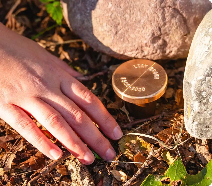 Close-up of a hand reaching toward a simple memorial marker along a rock-lined path in the Spreading Grove, symbolizing a peaceful resting place in nature.