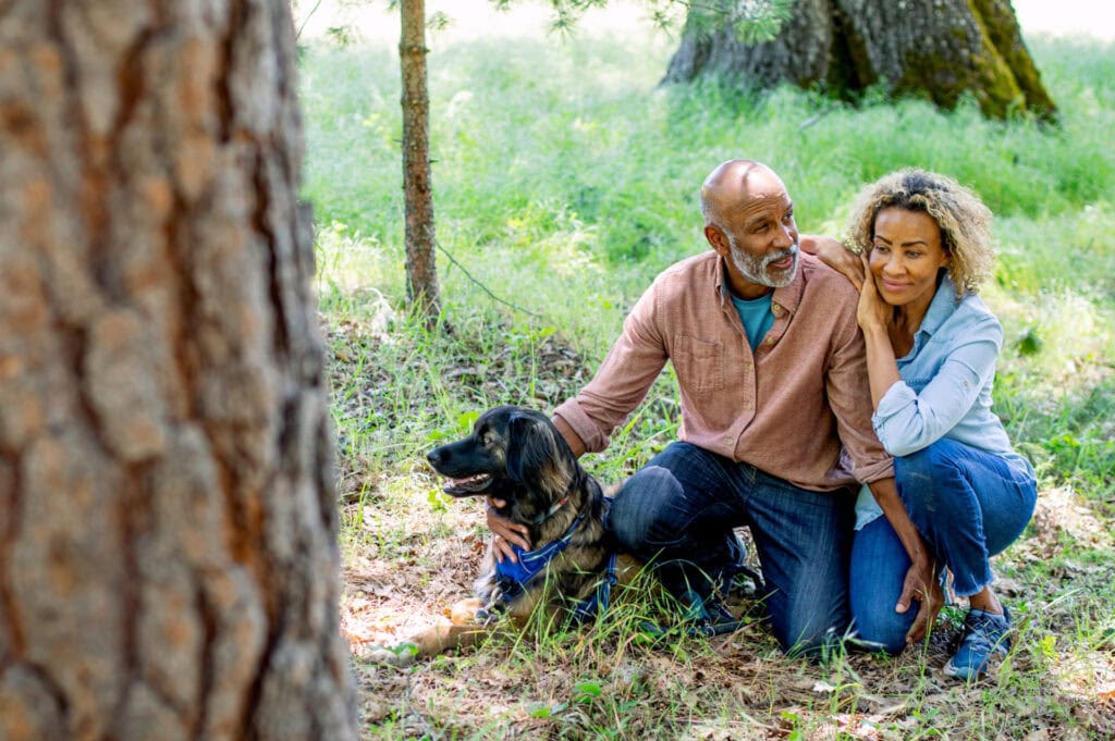 An adult couple kneeling together with their dog in a natural outdoor setting, observing the trees that surround them.