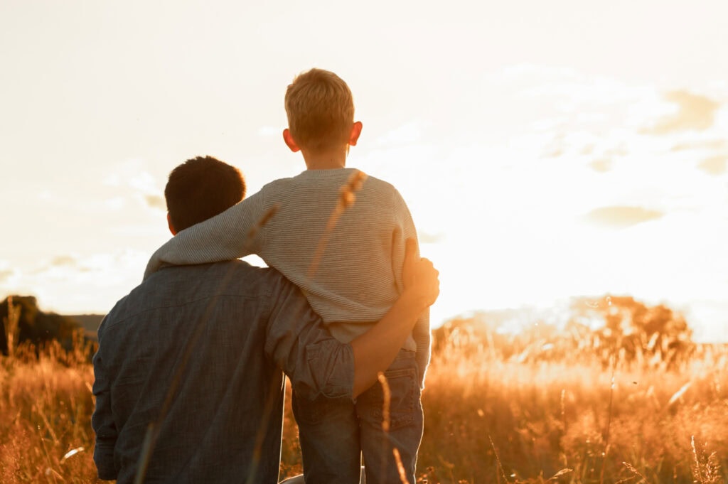 Back view of a parent sitting next to a child standing in a golden-lit field at sunset.