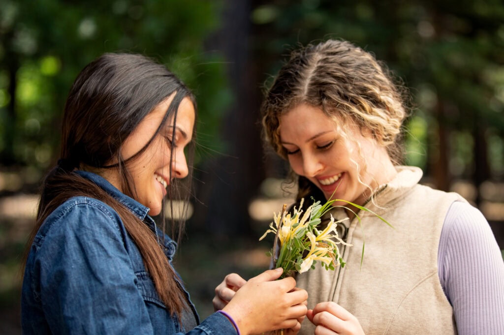 Two women standing close together outdoors, one holding white flowers, sharing a warm moment.