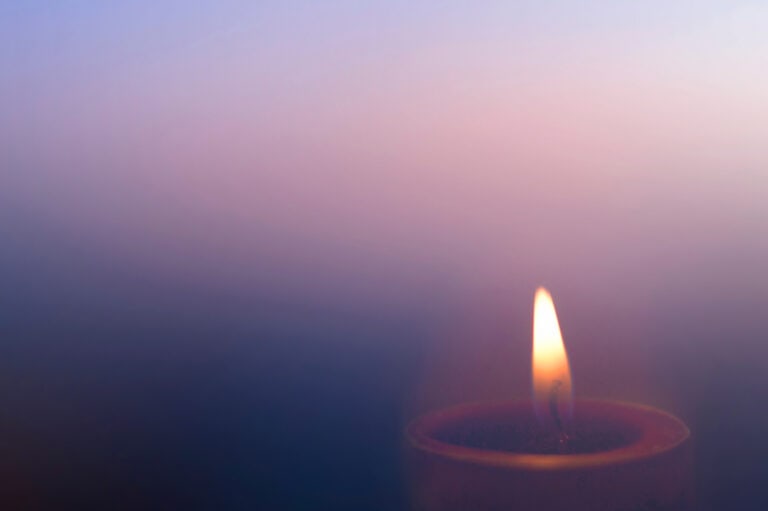 Single lit candle with a soft, glowing flame against a muted purple-blue background.