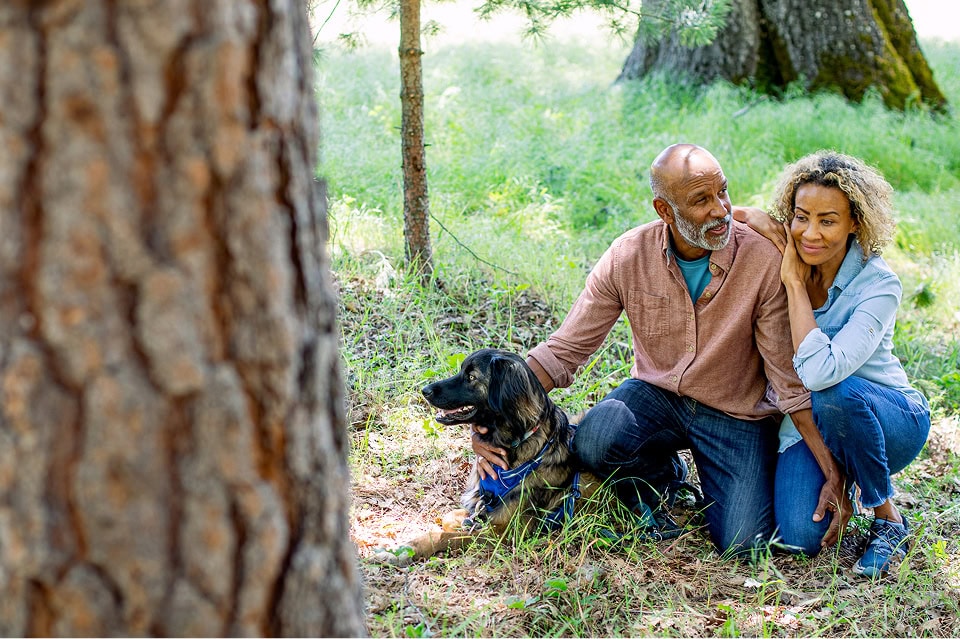 A middle-aged couple crouches in a forest clearing, smiling while petting their dog at the base of tree trunk in the forest.