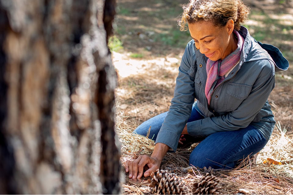A woman with curly dark hair crouches at a tree examining a Memorial Marker anchored in the ground at the tree's base.