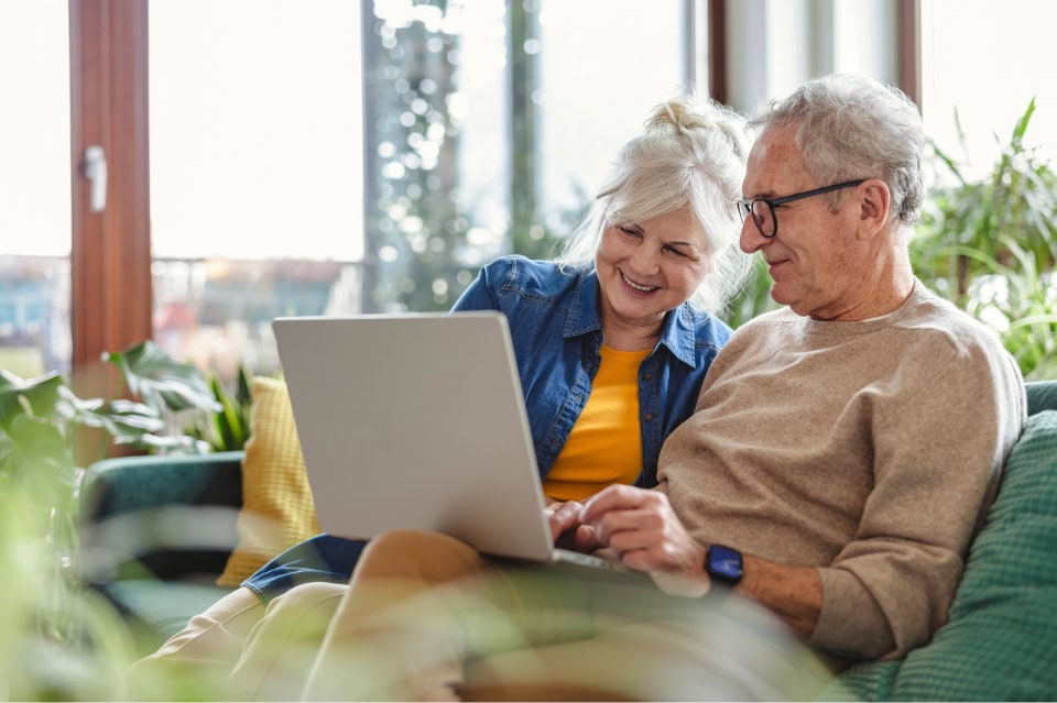 An older couple sits together on a couch, smiling and looking at a laptop.
