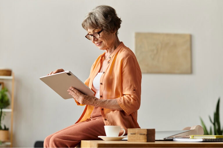 An older woman with gray hair and glasses sits on a desk, smiling while reading from a tablet.