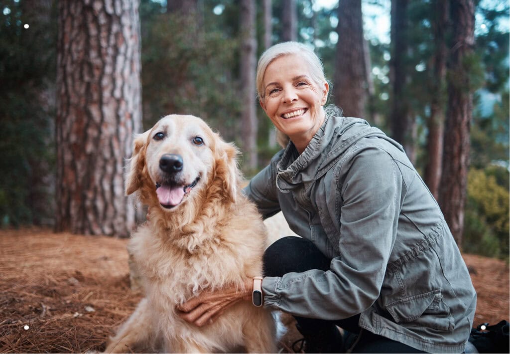 Smiling woman kneels beside her golden retriever in a forest setting.