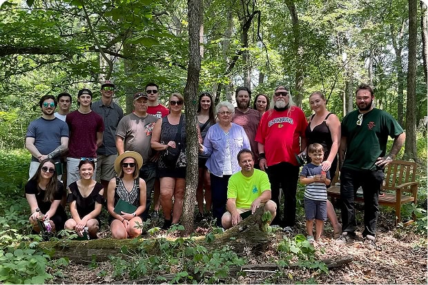A large multigenerational family group poses together outdoors in a wooded area, some seated and some standing, all smiling.
