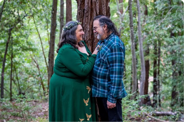 A bride and groom faces each other tenderly in a lush green forest, the woman wearing a floral crown and a green dress.
