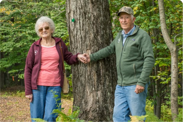 An older couple stands hand in hand beside a large tree in a forest, smiling gently at the camera.