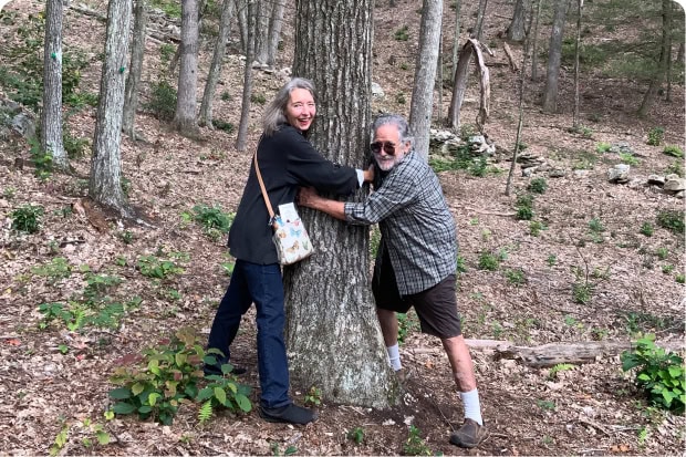 A couple embraces a large tree trunk in a woodland setting, both smiling and leaning into the tree.
