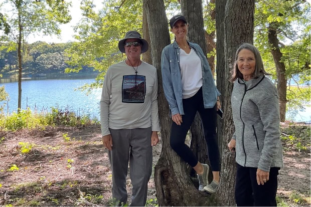 Three people stand smiling near a large tree with a lake visible in the background on a sunny day.