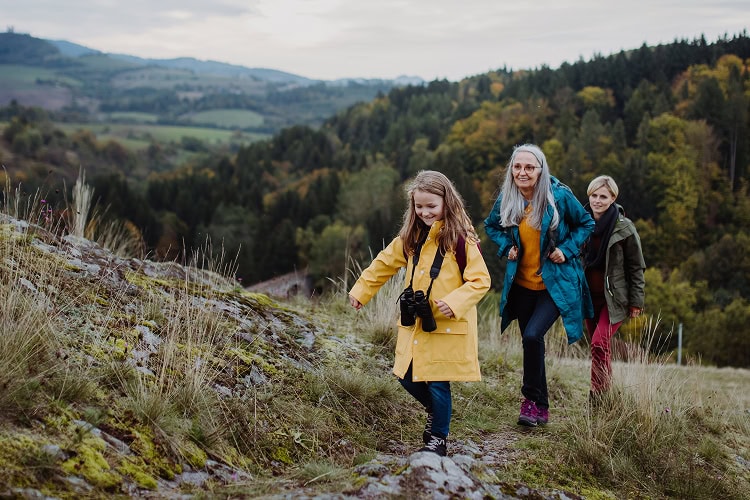 Three generations of women—grandmother, mother, and young granddaughter—hike up a grassy hill on a cloudy day with forest in the background.