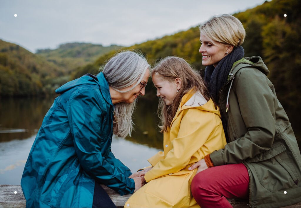 Three generations of women—grandmother, mother, and young granddaughter—share a joyful moment together on a bench overlooking the water.