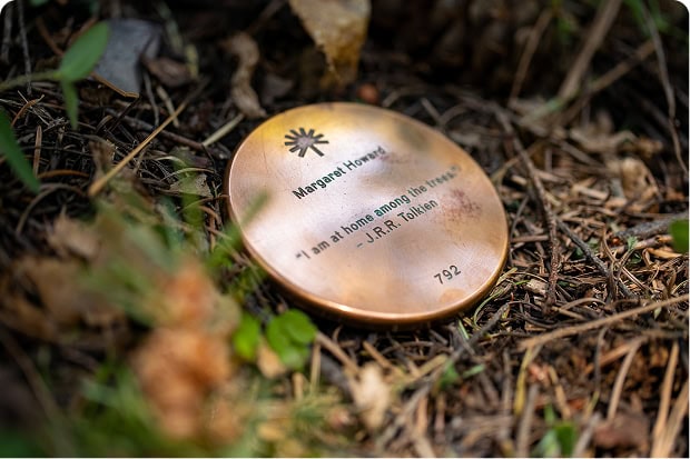 A closeup of round bronze memorial marker engraved with a name and a personal quote, anchored at the base of a Memorial Tree in the forest.