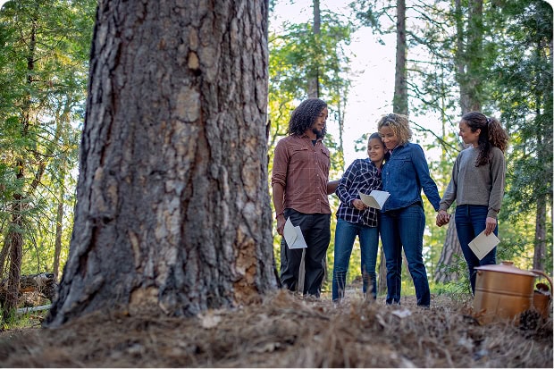 A family of four stands together near the base of a large tree in a forest during a Nature Memorial ceremony, sharing a tender moment.