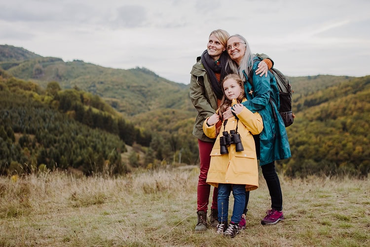 Three generations of women—grandmother, mother, and young granddaughter—standing at the top of a grassy hill embraced.