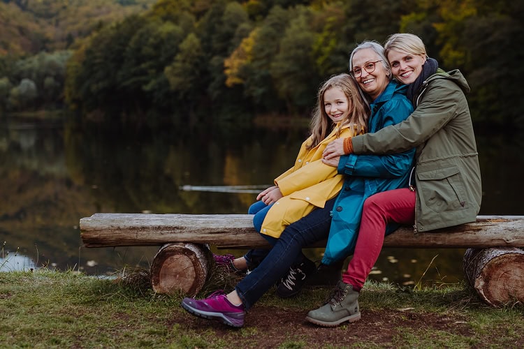 A grandmother, mother, and young daughter sitting together on a log bench by water, smiling and embracing in a forest setting.