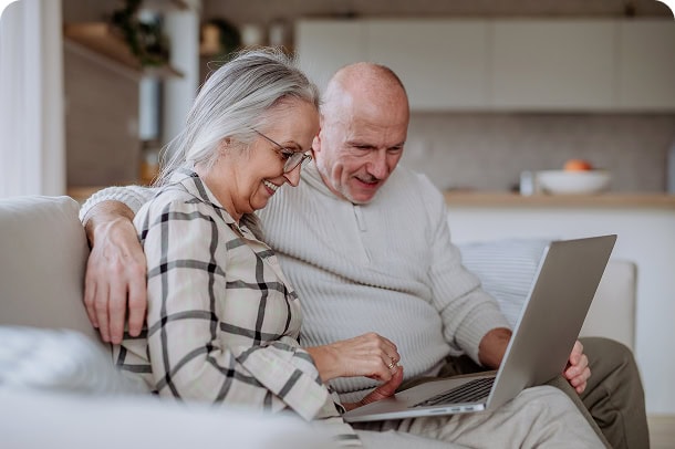 An older couple smiling together on a couch, looking at a laptop.