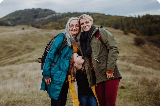 Three generations of women embracing outdoors in a a dry-grass hillside.