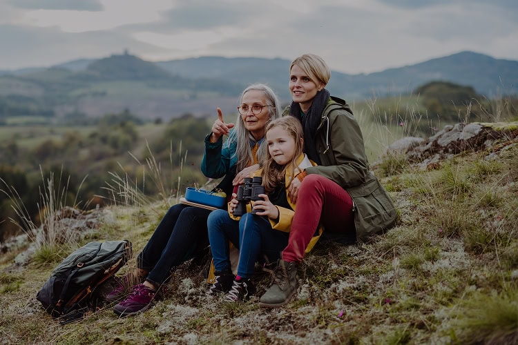 Three generations of women—grandmother, mother, and young granddaughter—sitting on a grassy hillside looking at something in the distance.