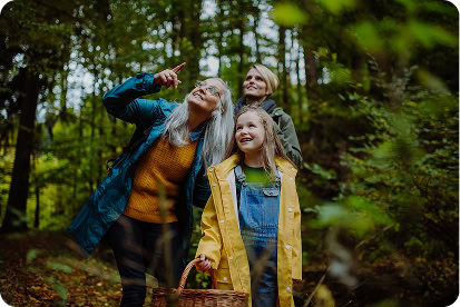 Three generations of women—grandmother, mother, and young granddaughter—stand together looking up at a Memorial Tree in the forest.