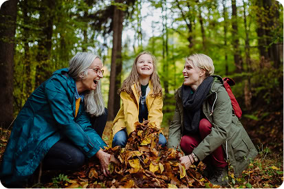 Three generations of women—grandmother, mother, and young granddaughter—play with a pile of leaves in the forest.