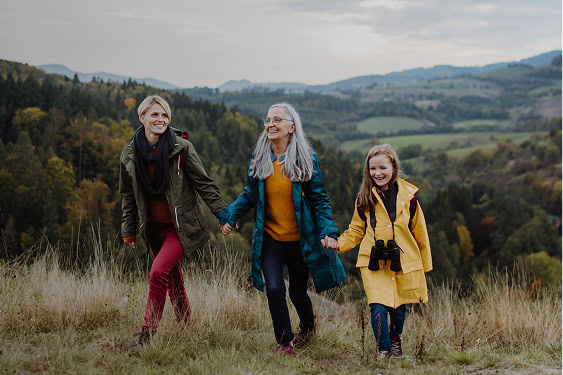 Three generations of women—grandmother, mother, and young granddaughter—walk uphill hand in hand, with rolling hills in the background.