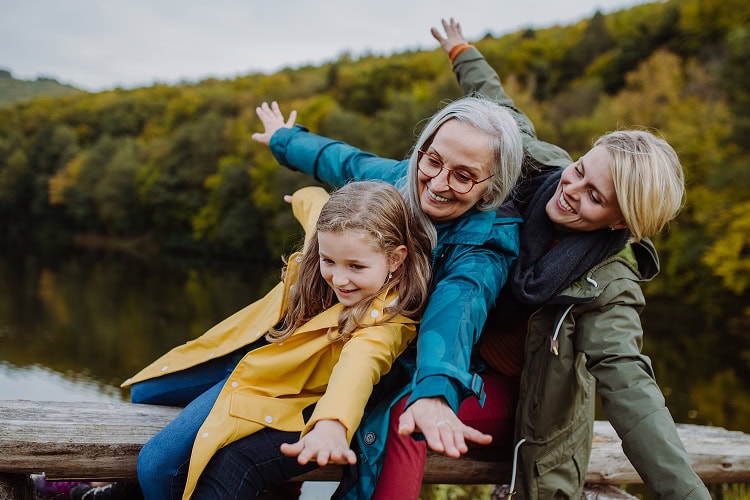 Three generations of women—grandmother, mother, and young granddaughter—sitting on a log bench by water, tilting to one side smiling.