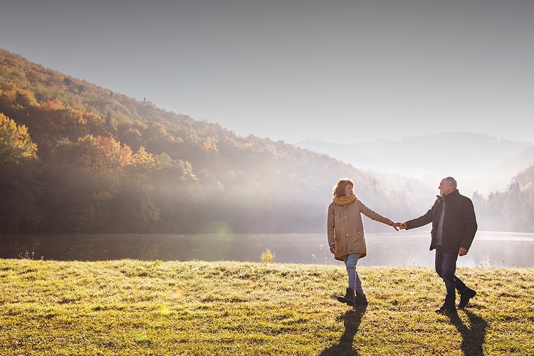 A couple holding hands on the grass in front of a body of water on a sunny day with rolling hills in the background.