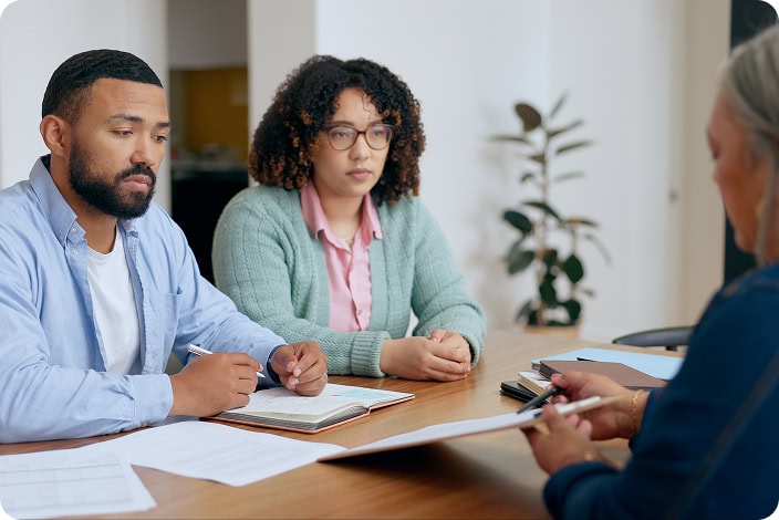 An unhappy looking younger couple sitting across a table from a person in a meeting discussing paperwork.