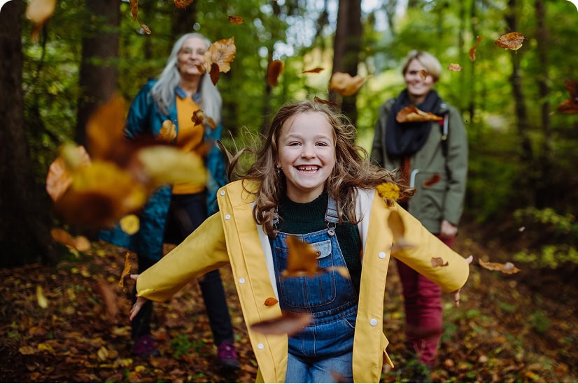 A young girl in a yellow raincoat laughs joyfully as leaves swirl around her; her mother and grandmother are smiling in the background.
