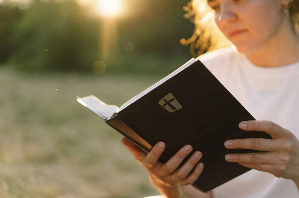 A young woman in a white t-shirt reading the Bible outdoors at sunset.