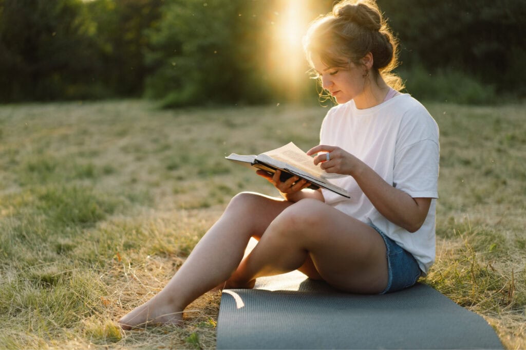 A young woman in a white t-shirt sitting down outside reading the Bible at sunset.