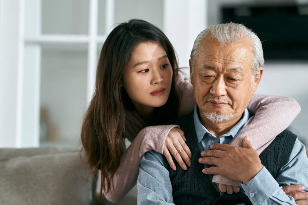 A younger woman resting her head on an older man's shoulder, both looking somber.