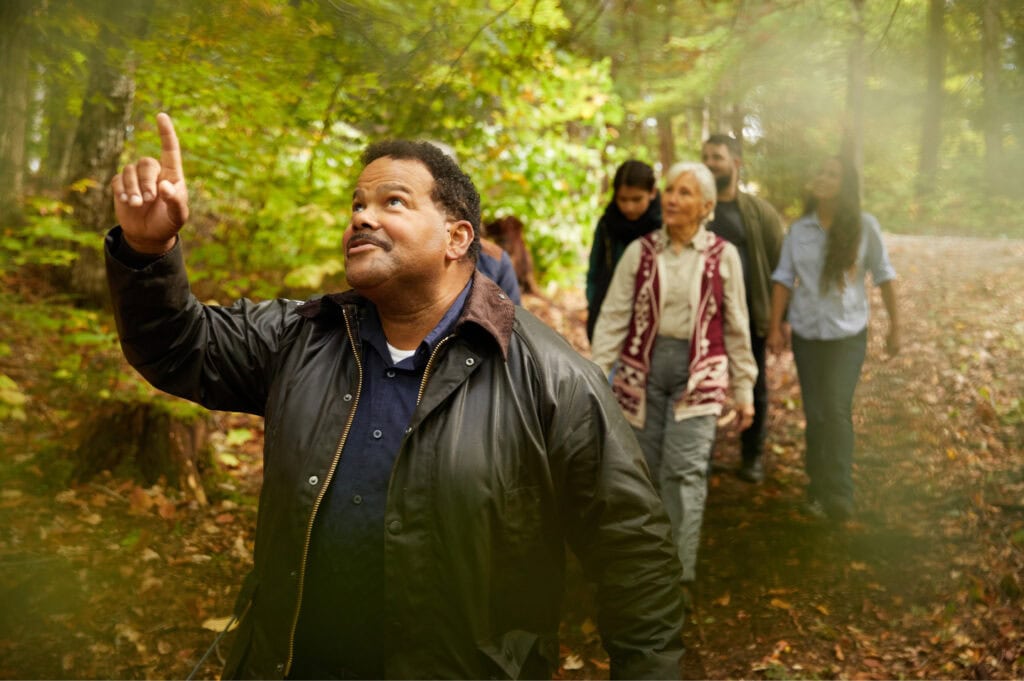 A man points upward toward the tree canopy on a forest path, with a group of people walking behind him.