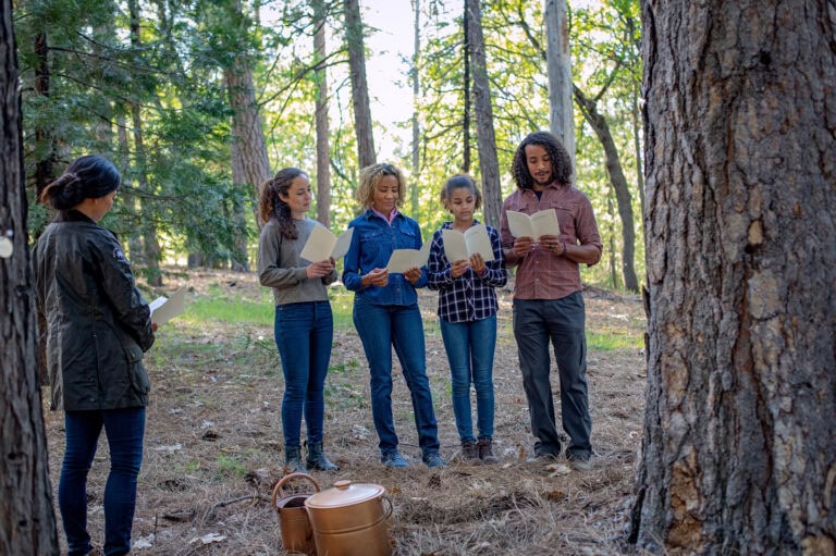 A small group of people standing among trees in a forest, each holding a booklet during an outdoor memorial ceremony.