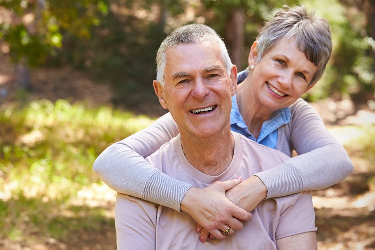 A smiling older couple in a sunlit forest with the woman embracing the man from behind.