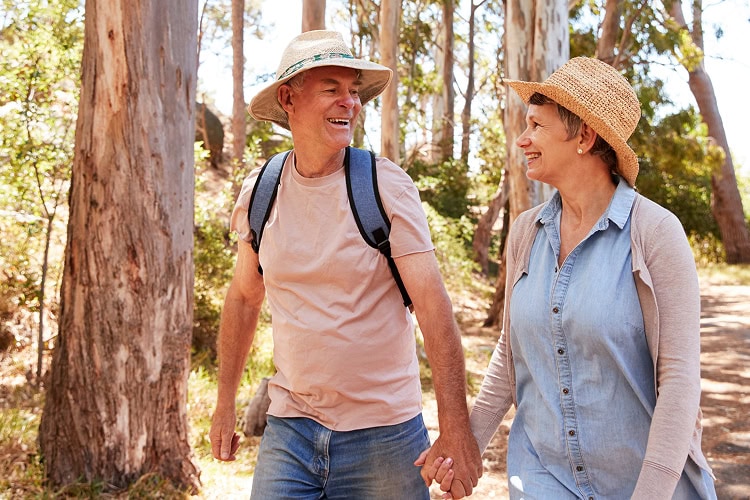 An older couple walking hand in hand in a sunlit forest smiling at each other.