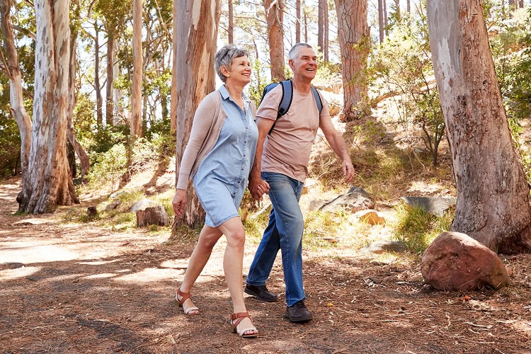 A smiling older couple in a sunlit forest walking hand in hand.
