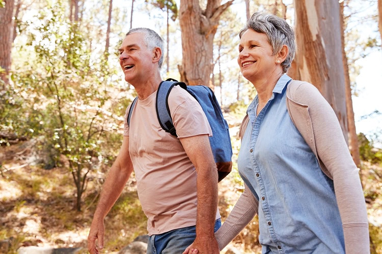 A older couple walking hand in hand in a sunlit forest smiling at each other.