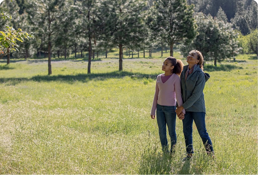 A mother and daughter holding hands, walking through a sunlit meadow, surrounded by trees.