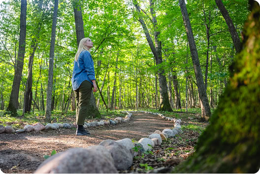 A woman standing in a lush green forest along a path lined with stones, surrounded by tall trees.