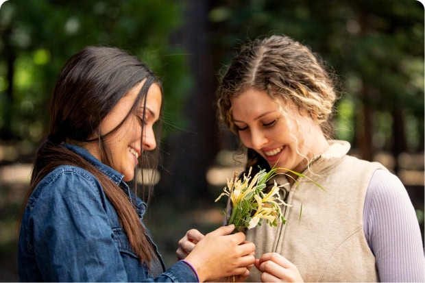 Two adult sisters smiling at each other in the forest, one holding a small bunch of wildflowers.