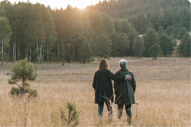 The back view of a woman and her mother in a dry grassy meadow looking at the sunset in the forest in the distance.