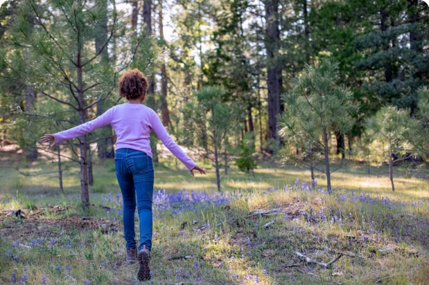 A child running with arms outstretched through an open forest meadow with purple wildflowers.