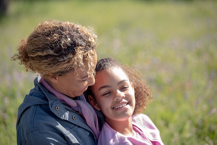 A mother lovingly embraces her young daughter with sunlight illuminating their smiling faces.