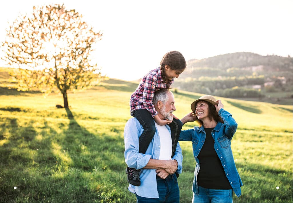 Grandparents and granddaughter enjoying a sunny day in a green field, with the young girl riding on her grandfather's shoulders.
